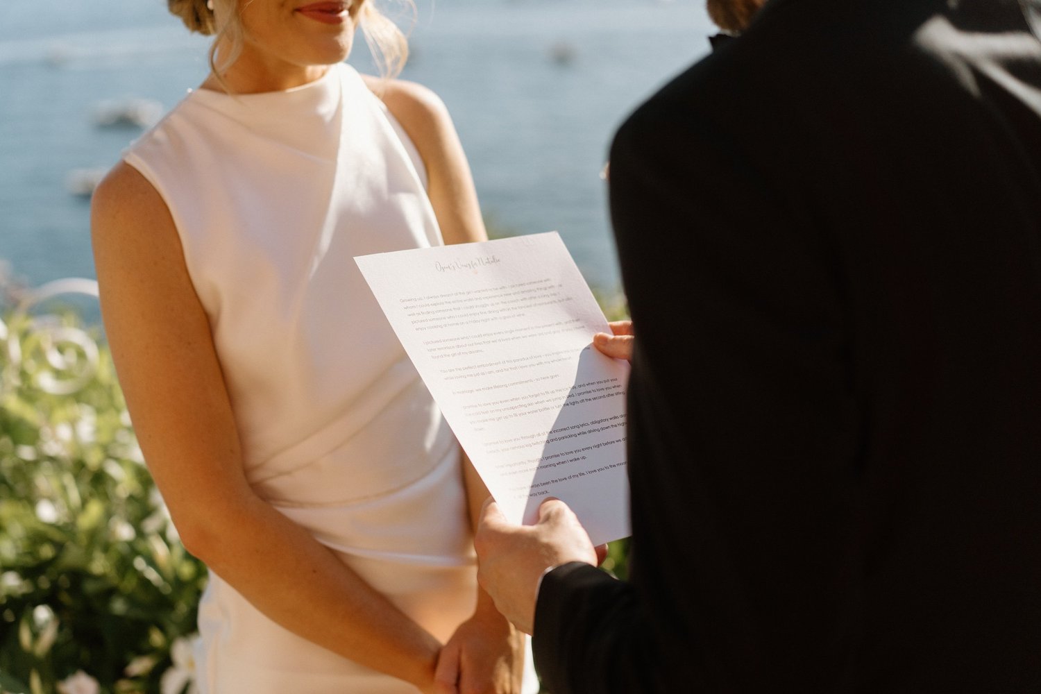A person in a black jacket holds a sheet of paper during a ceremony, with a woman in a sleeveless white dress smiling and standing outdoors near water and greenery.