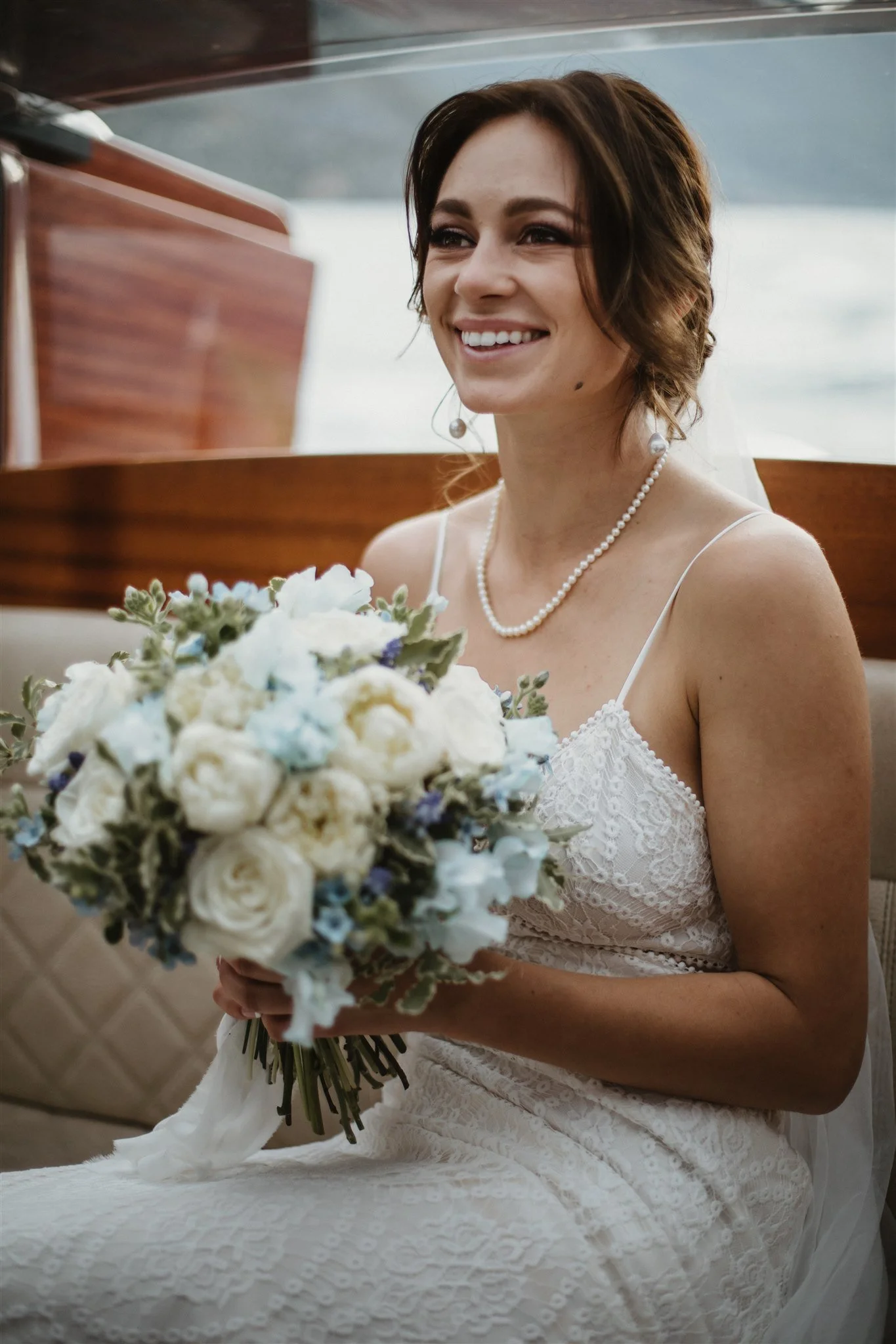 A smiling bride in a white lace wedding dress holding a bouquet of white and blue flowers, wearing pearl jewelry, sitting in a boat near water.