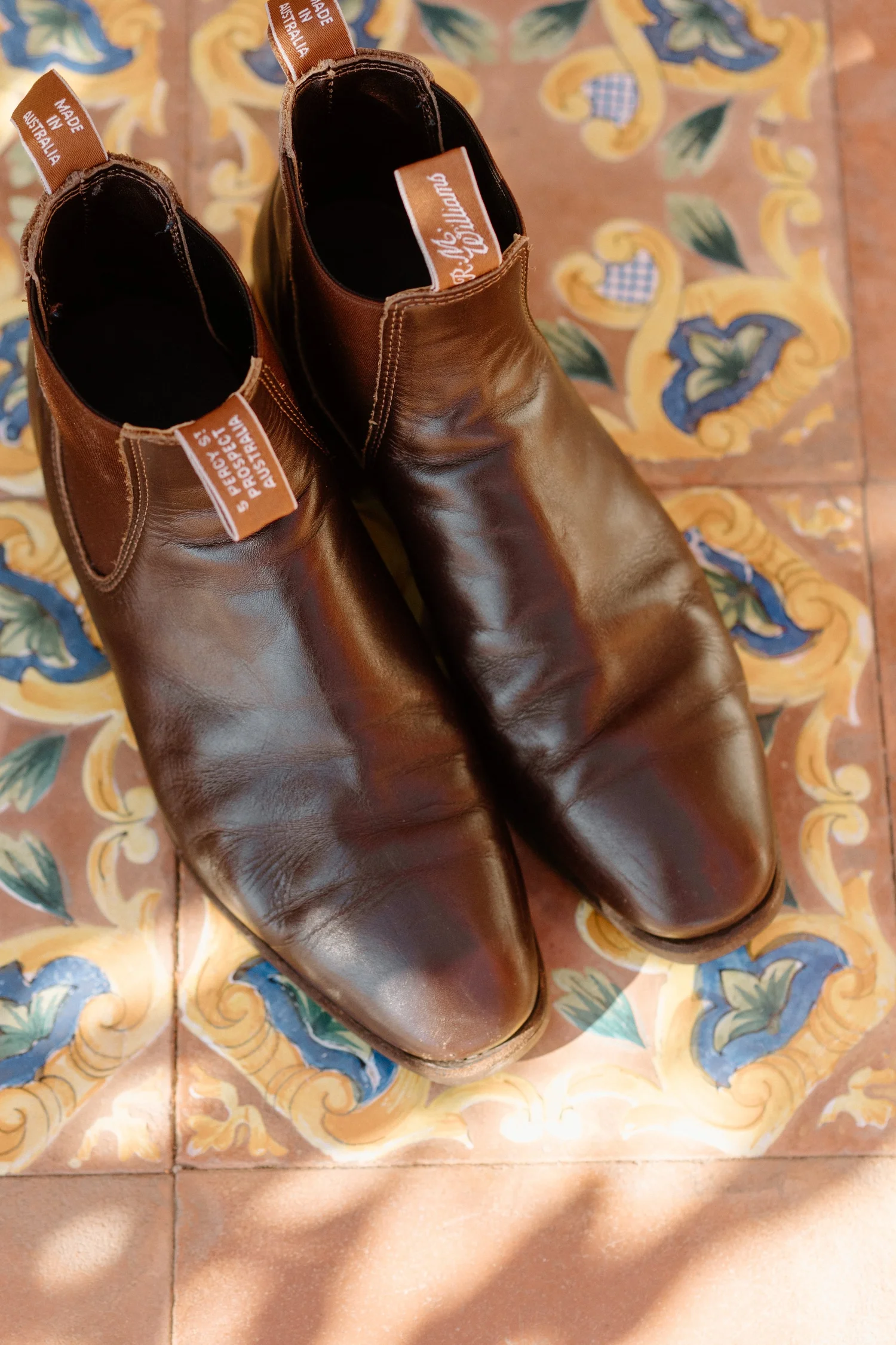 A pair of brown leather boots with tags that read 'Made in Australia' are placed on a decorative tiled floor with floral patterns in yellow, blue, green, and peach colors.