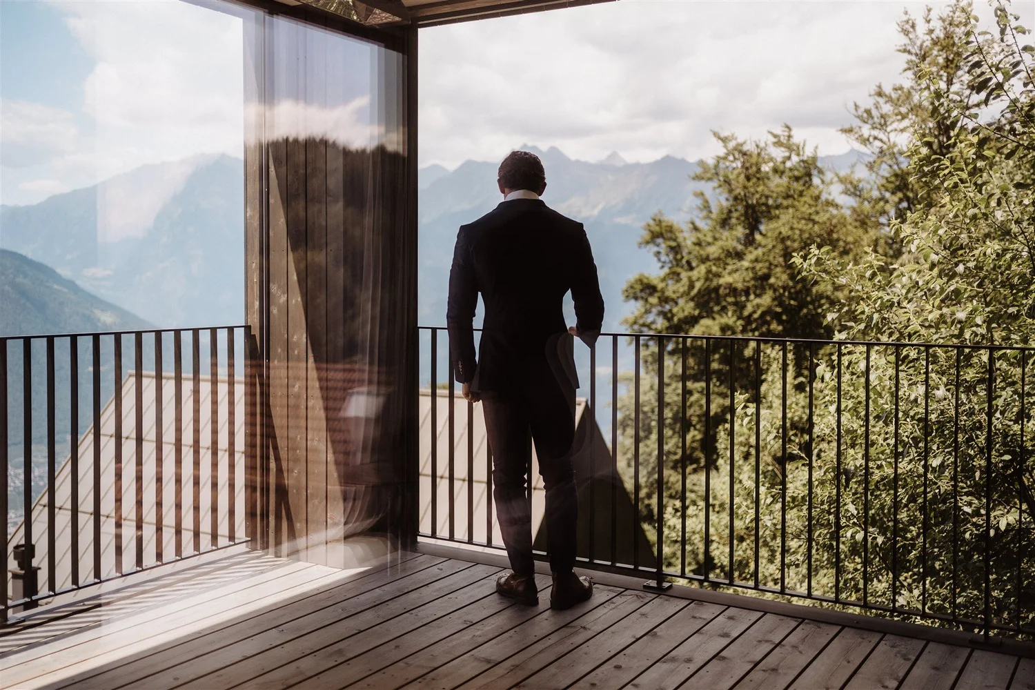 A man in a suit standing on a wooden balcony, looking out at a mountainous landscape with trees and clouds.