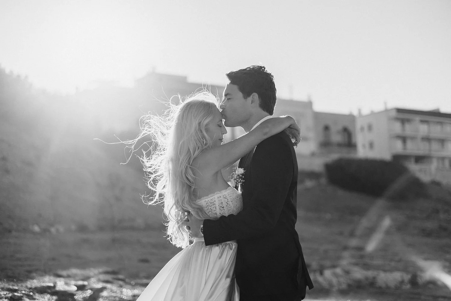 A black-and-white photo of a couple embracing on a beach, with the woman resting her head on the man's shoulder and the man kissing her forehead.