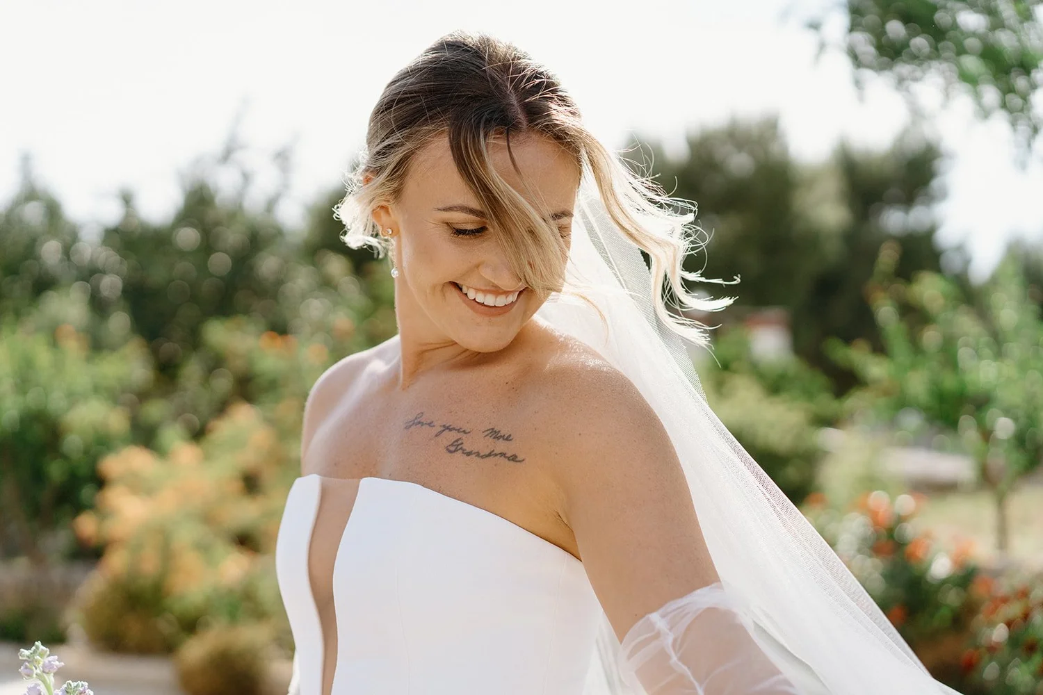 A smiling woman in a white wedding dress with a veil outdoors, looking down at her shoulder, with a tattoo that reads 'Love you more Grandma'.