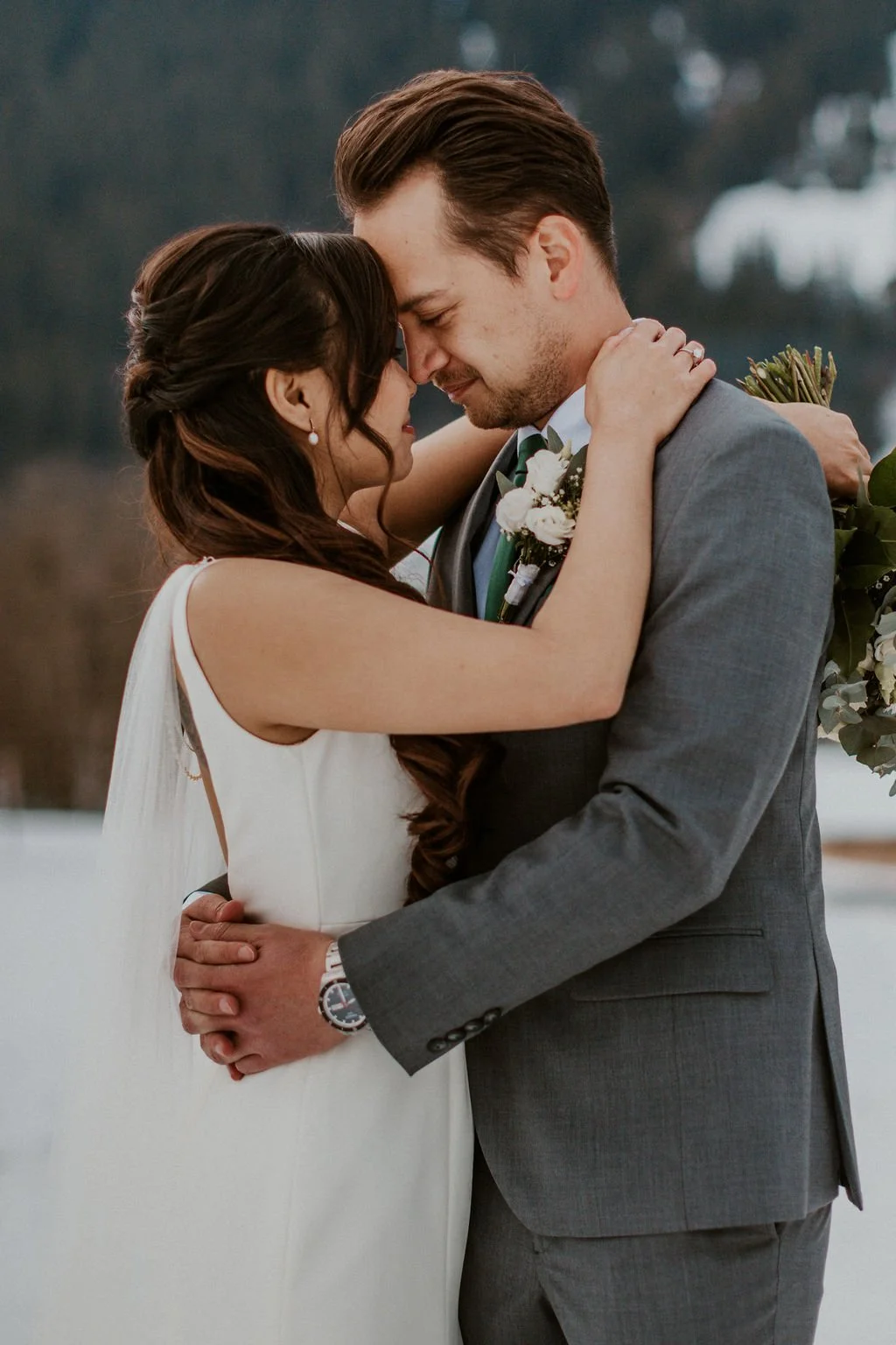 A bride and groom embrace during their wedding ceremony outdoors in winter. The bride has long brown hair and wears a white dress, and the groom has short brown hair, a beard, and wears a gray suit with a boutonniere. They hold each other's faces wit