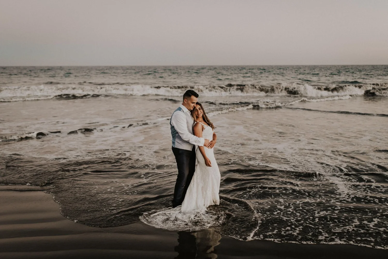 A couple in wedding attire standing closely on the shoreline with ocean waves in the background during sunset.