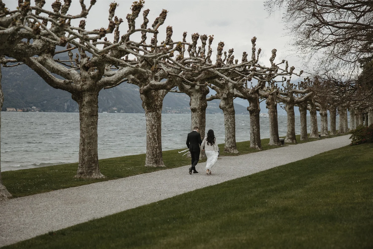 A man and woman dressed in wedding attire walking along a lakeside park path. The scene is cloudy and overcast, with leafless trees lining the walkway and mountains in the background.