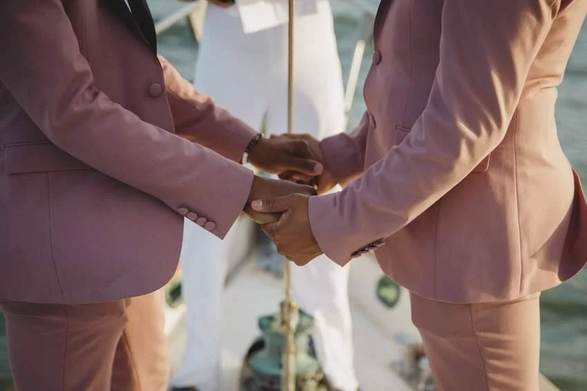 Two people holding hands during a wedding ceremony on a boat, dressed in pink suits.