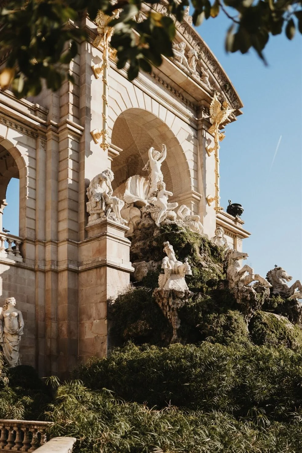 Statues and sculptures on a historic building's facade, with a large arched opening, ornate decorations, and golden accents, under a clear blue sky with some greenery in the foreground.