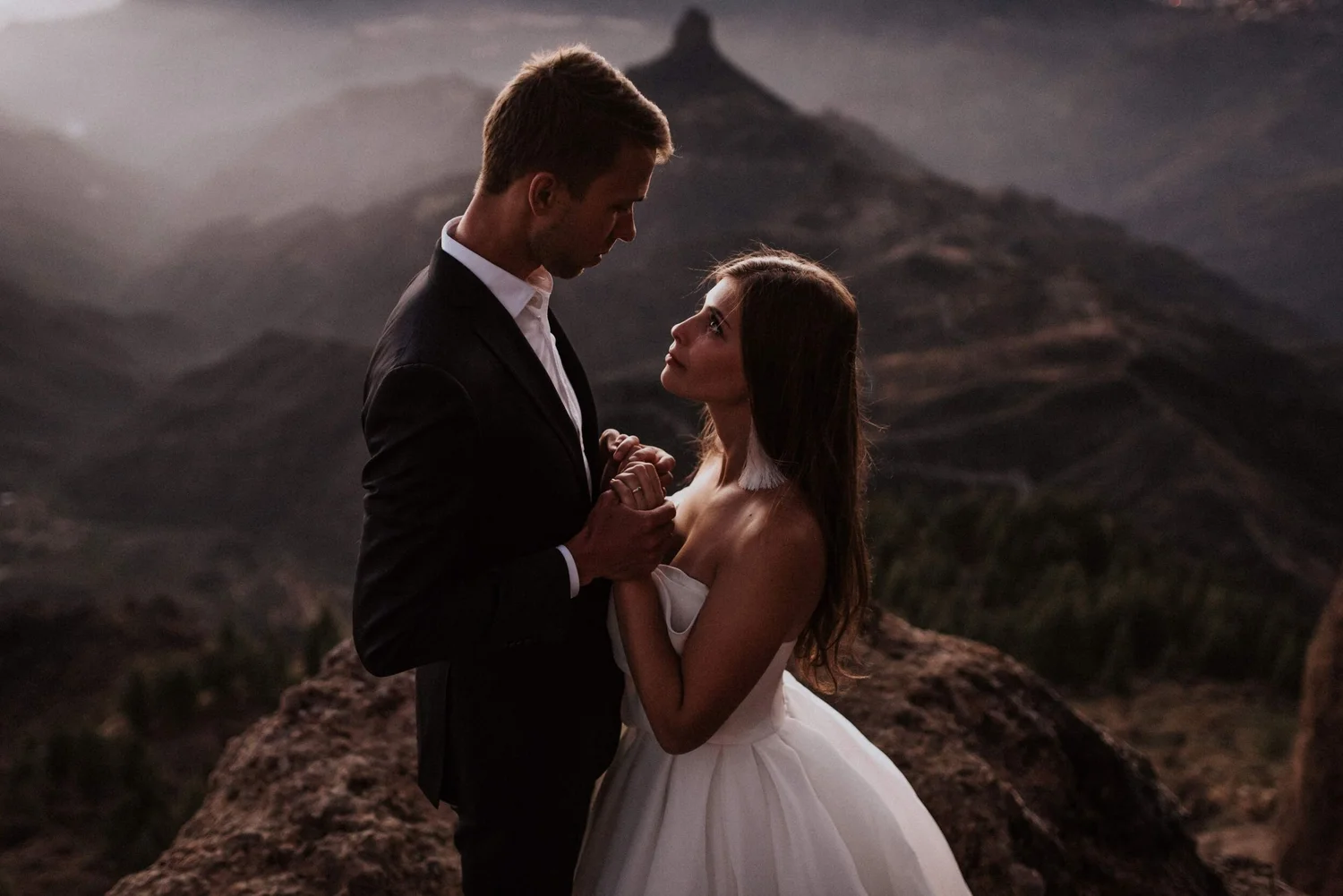 A man in a black suit and woman in a white dress holding hands and looking at each other outdoors with mountains in the background during sunset.