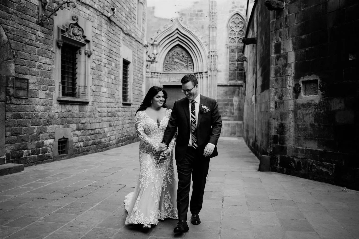 A black and white photo of a wedding couple walking hand in hand along a narrow, historic street with stone walls and arched doorway in the background.