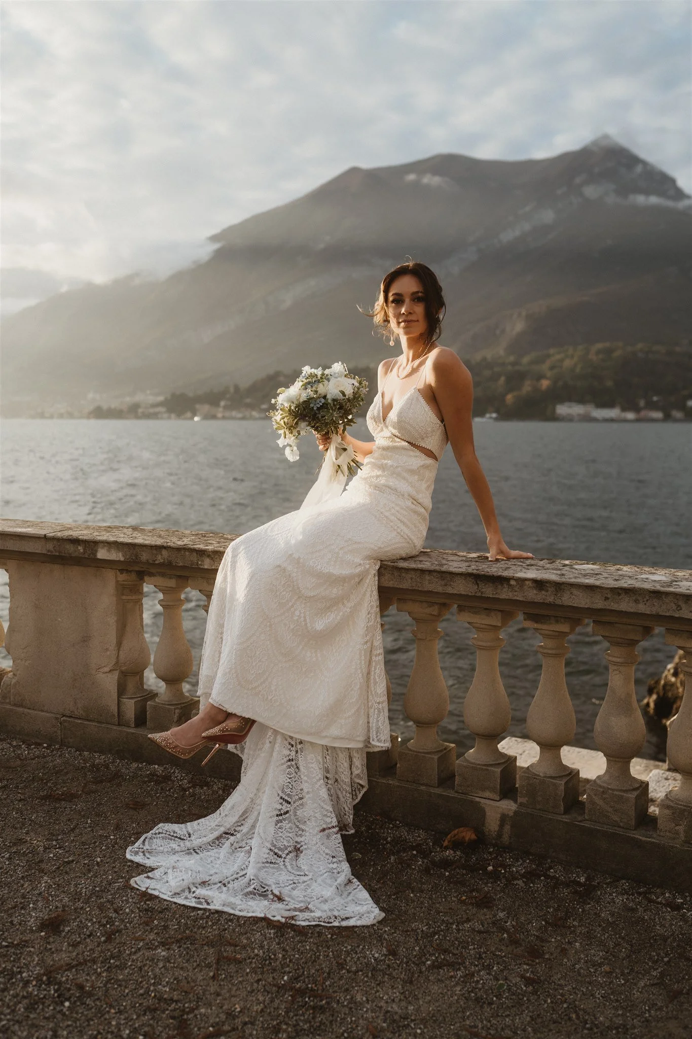 A woman in a lace wedding dress sitting on a stone railing near a body of water with mountains in the background at sunset.