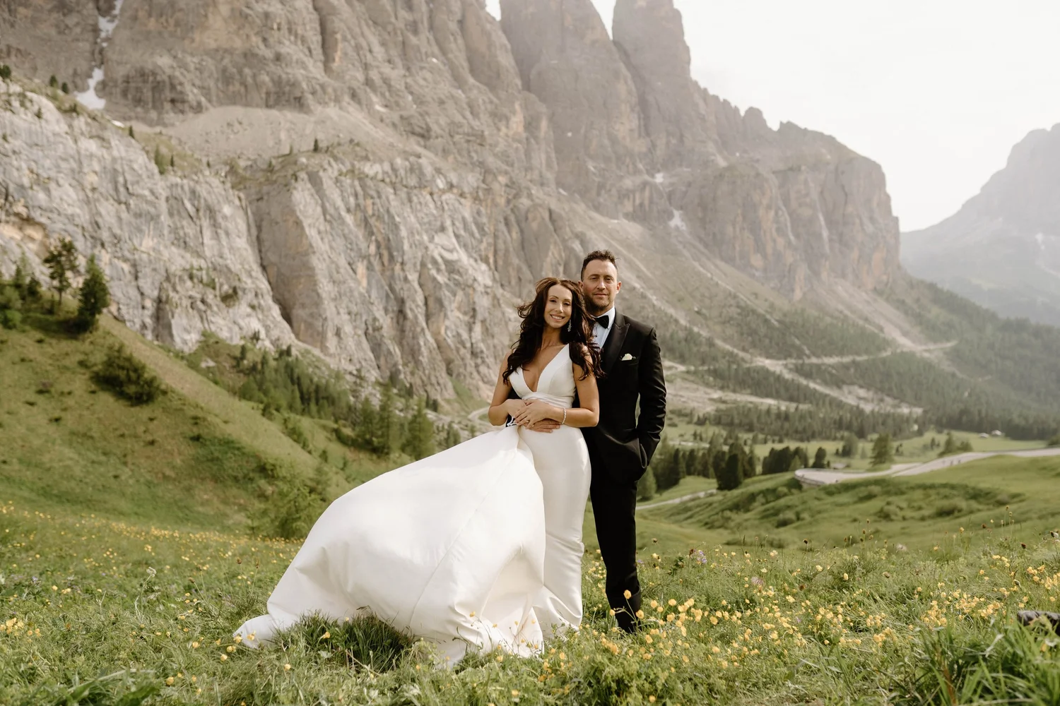 A bride and groom stand together in a grassy field with yellow flowers, mountains and a cloudy sky in the background.