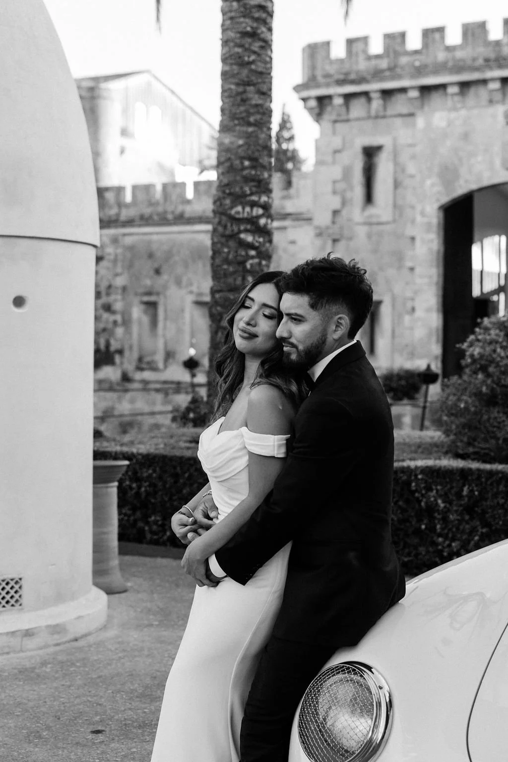 A black and white photo of a couple dressed in formal attire, leaning against a vintage car. The woman has long wavy hair and wears an off-shoulder dress, with her eyes closed and a gentle smile. The man has short hair and a beard, dressed in a tuxed