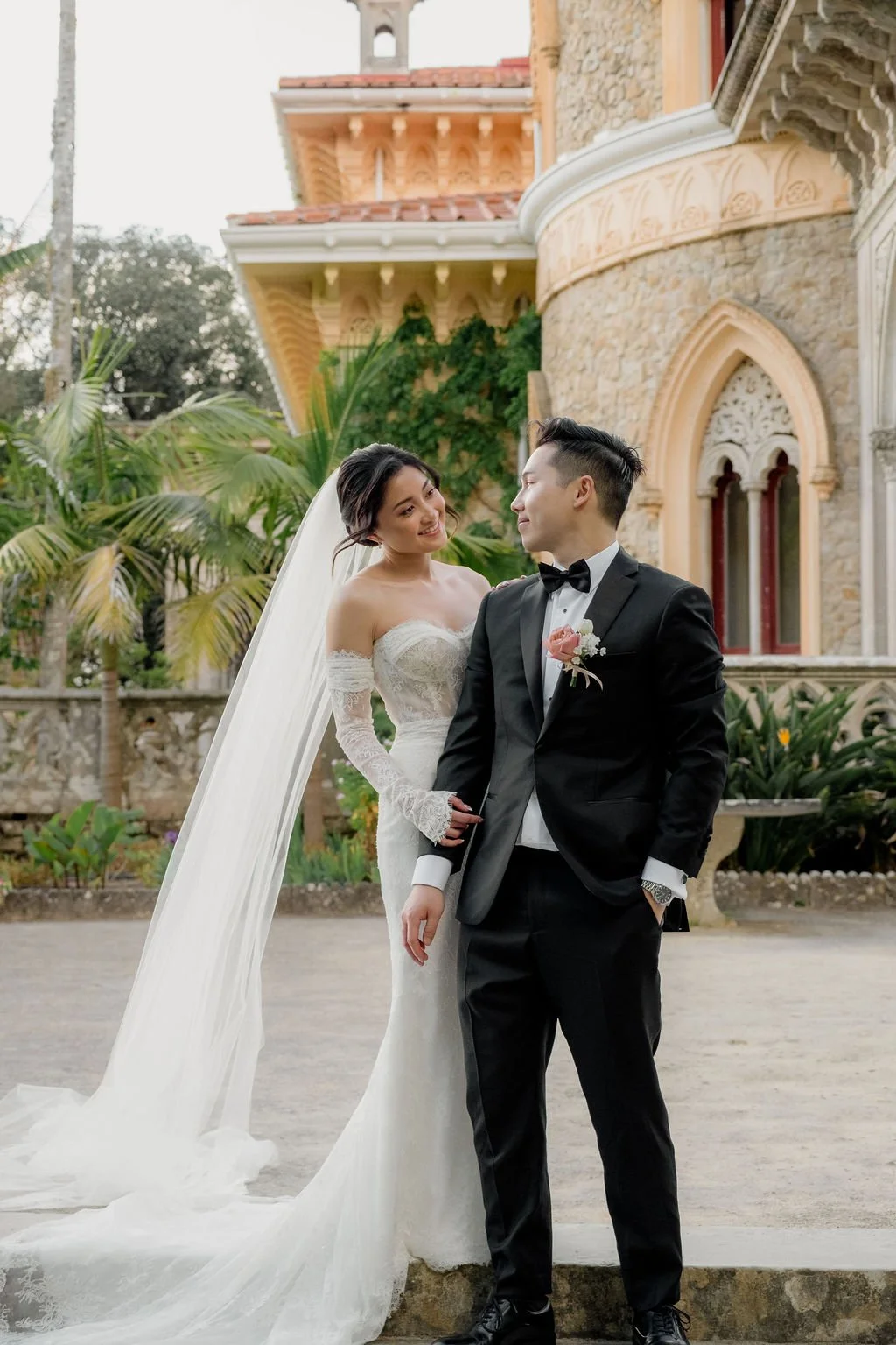 A bride in a white wedding gown and veil, and a groom in a black tuxedo with a pink boutonniere, stand outdoors in front of a historic stone building with arched windows and lush greenery.