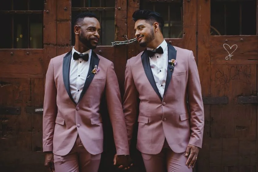 Two men in matching pink tuxedos and black bow ties holding hands and smiling at each other in front of a wooden barn door.