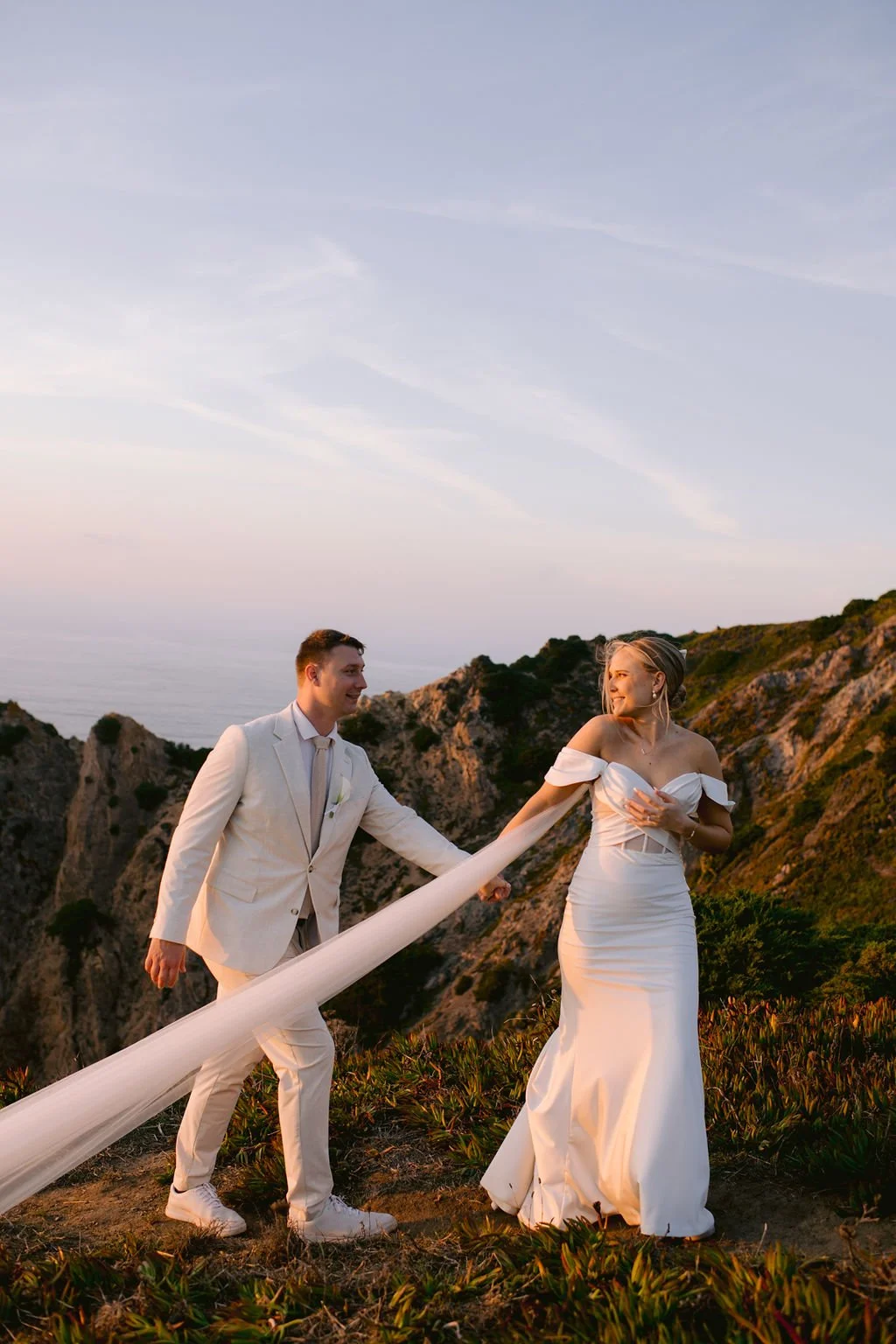 A bride and groom in wedding attire walking hand-in-hand outdoors on a hillside during sunset, with rocky cliffs and the ocean in the background.