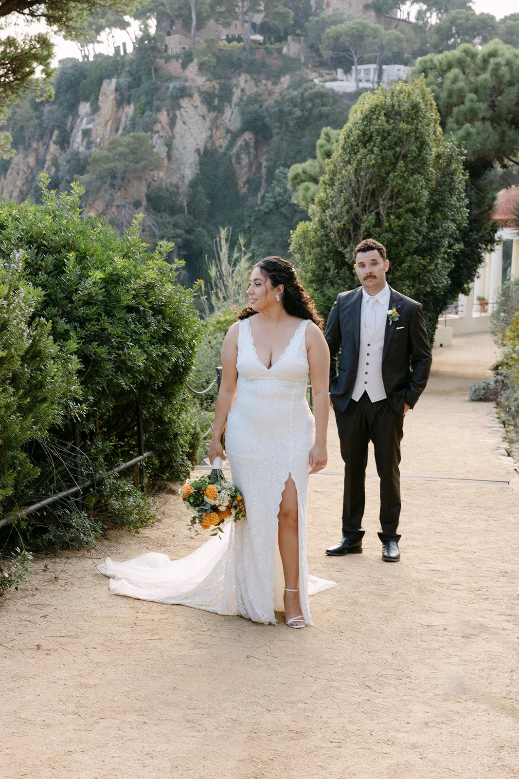 A bride in a white wedding dress holding a bouquet, and a groom in a black suit with a vest, walking on a garden path surrounded by greenery with hills in the background.