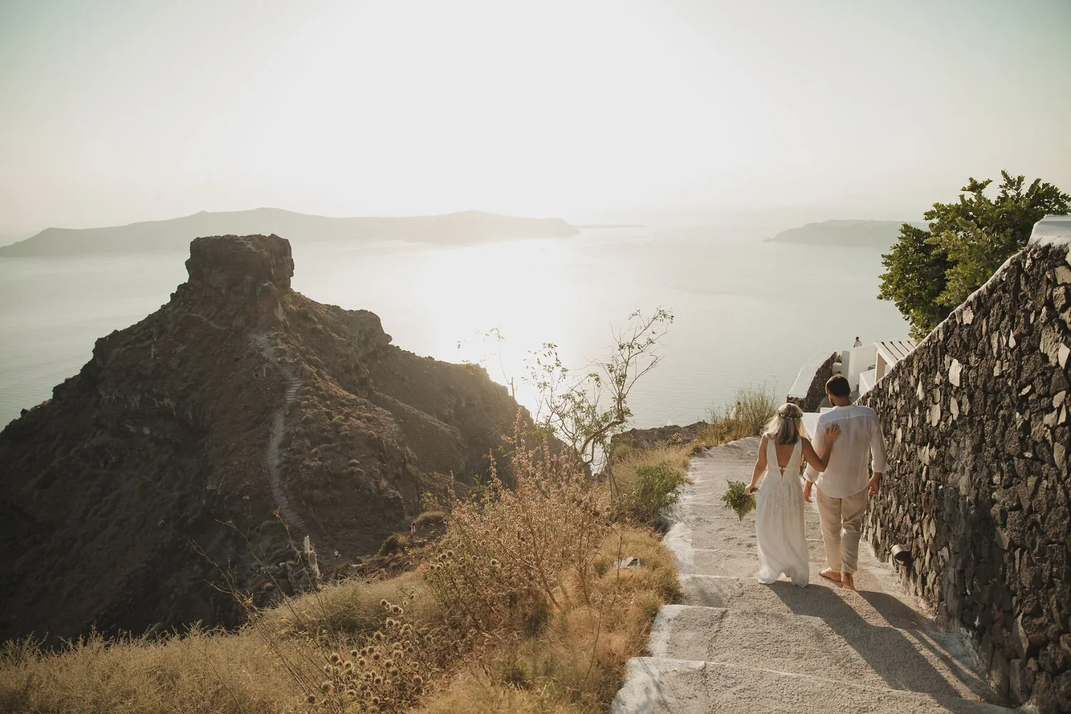 Couple walking hand in hand along a path overlooking a scenic coastal view with rocky cliffs and calm water, in a sunny setting.