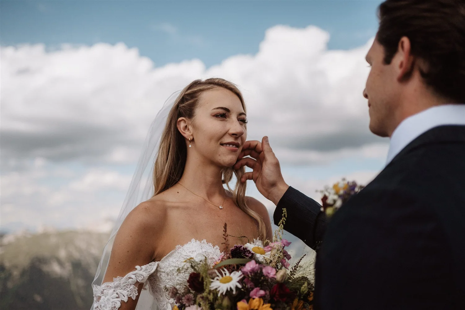 A bride and groom exchanging vows outdoors during the daytime, with the bride holding a bouquet of wildflowers, under a partly cloudy sky.