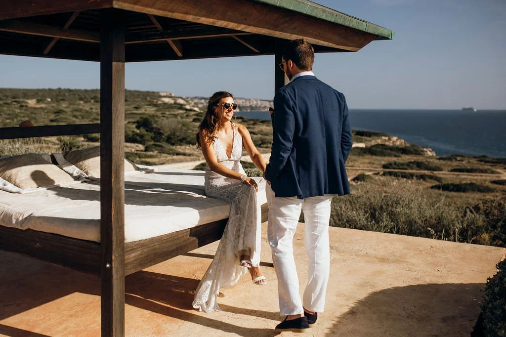 A woman in a white dress sitting on a bed outside, smiling and wearing sunglasses, while a man in a blue blazer and white pants stands nearby, with a scenic coastal landscape in the background.