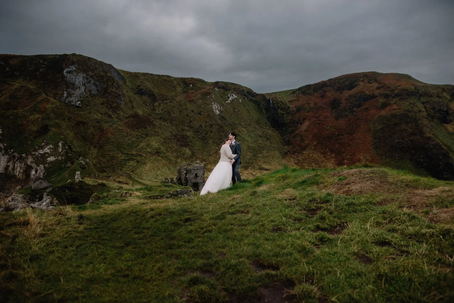 A bride and groom standing together outdoors against a mountainous landscape with a cloudy sky.