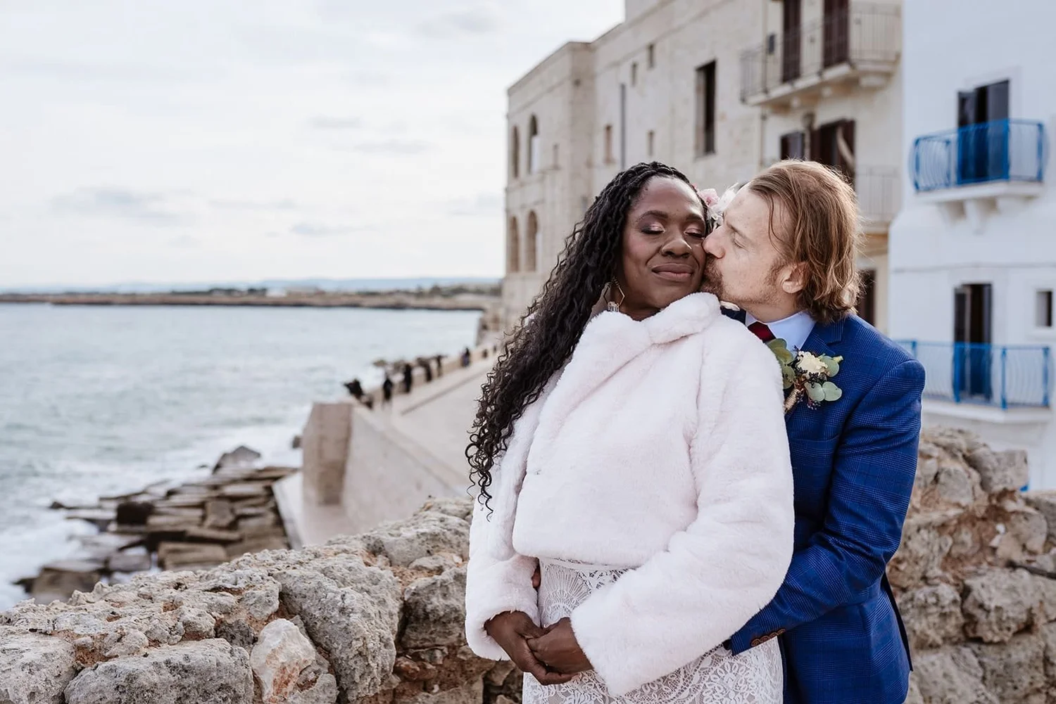 A couple dressed in wedding attire embracing by the water with a stone wall and buildings with balconies in the background, on a cloudy day.