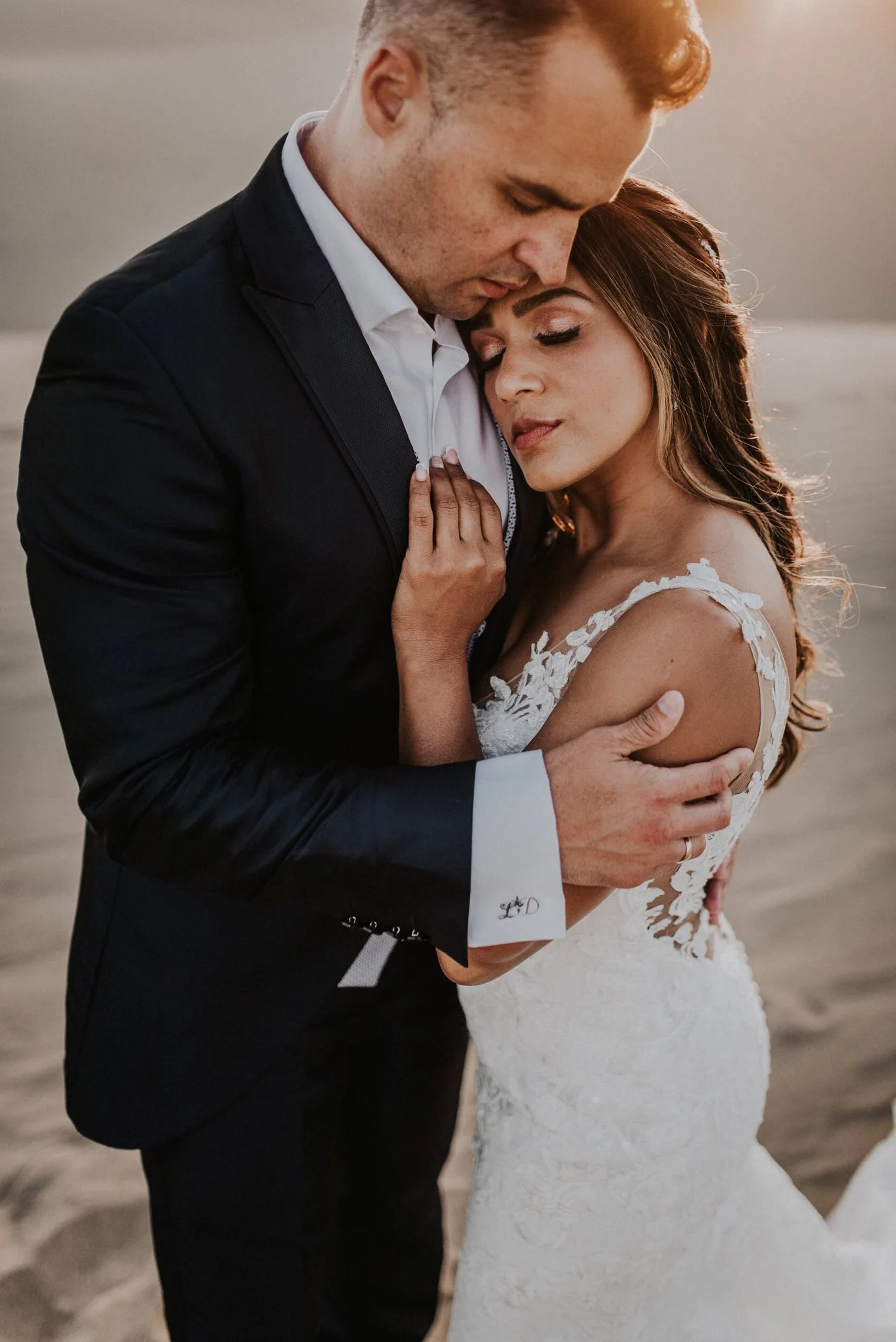 A couple in formal attire embracing at the beach during sunset, with the woman wearing a white lace wedding dress and the man in a black suit.