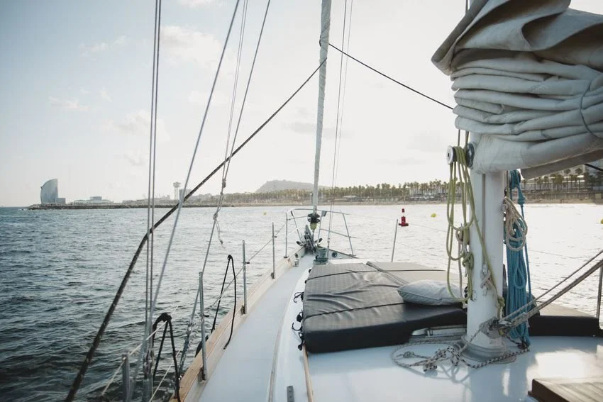 View from a sailboat's deck looking towards the water with a city skyline in the distance, including a distinctive sail-shaped building, under partly cloudy skies.