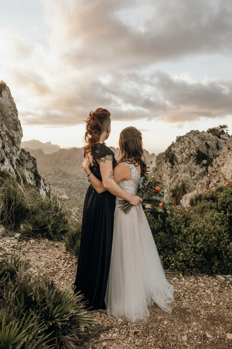 Two women in wedding dresses stand close together outdoors on rocky terrain with mountains and cloudy sky in the background during sunset.