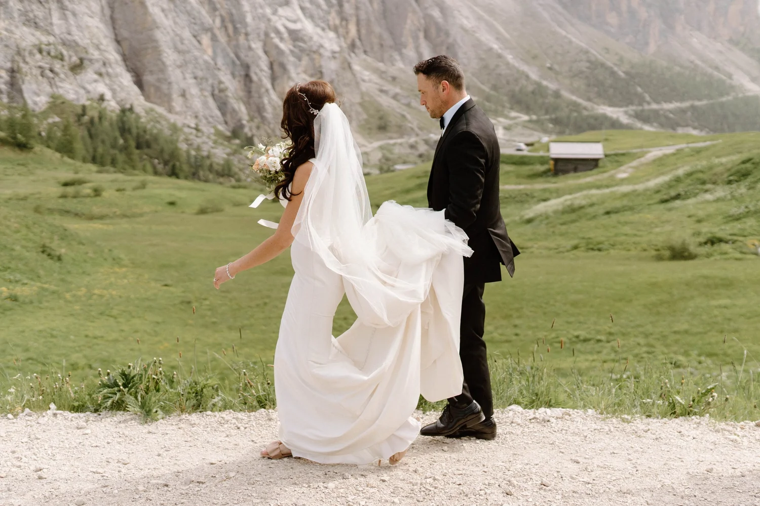 A bride and groom standing outdoors on a dirt path, with the bride holding a bouquet of flowers and wearing a wedding dress and veil, and the groom in a black tuxedo, in a scenic mountainous landscape.