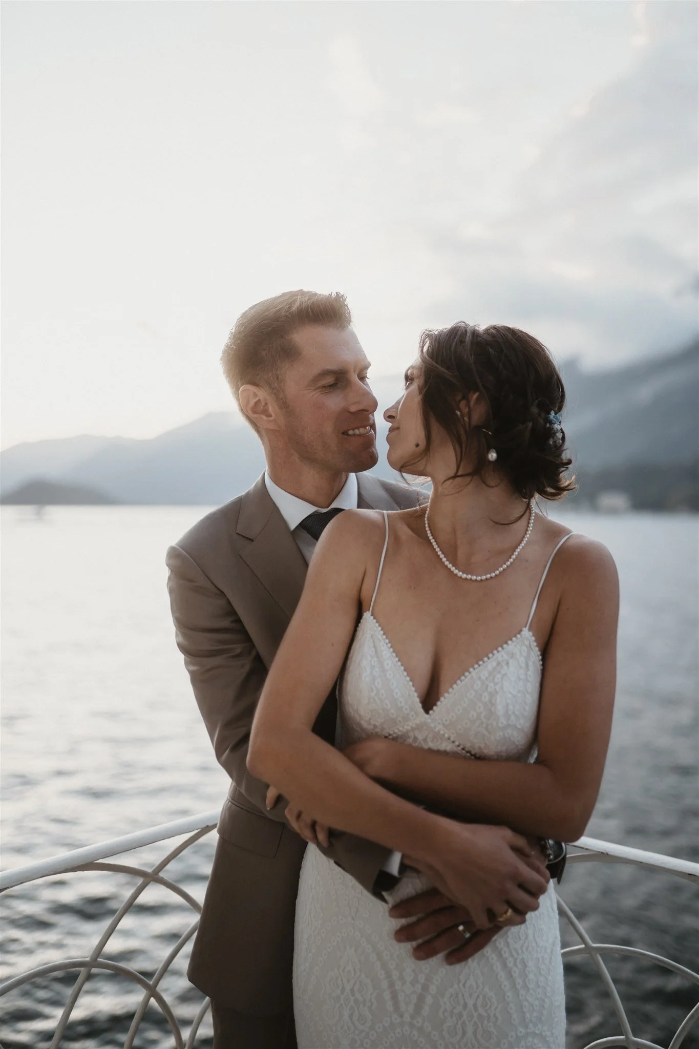 A couple dressed in wedding attire standing on a boat near a body of water, with mountains in the background, sharing an intimate moment.
