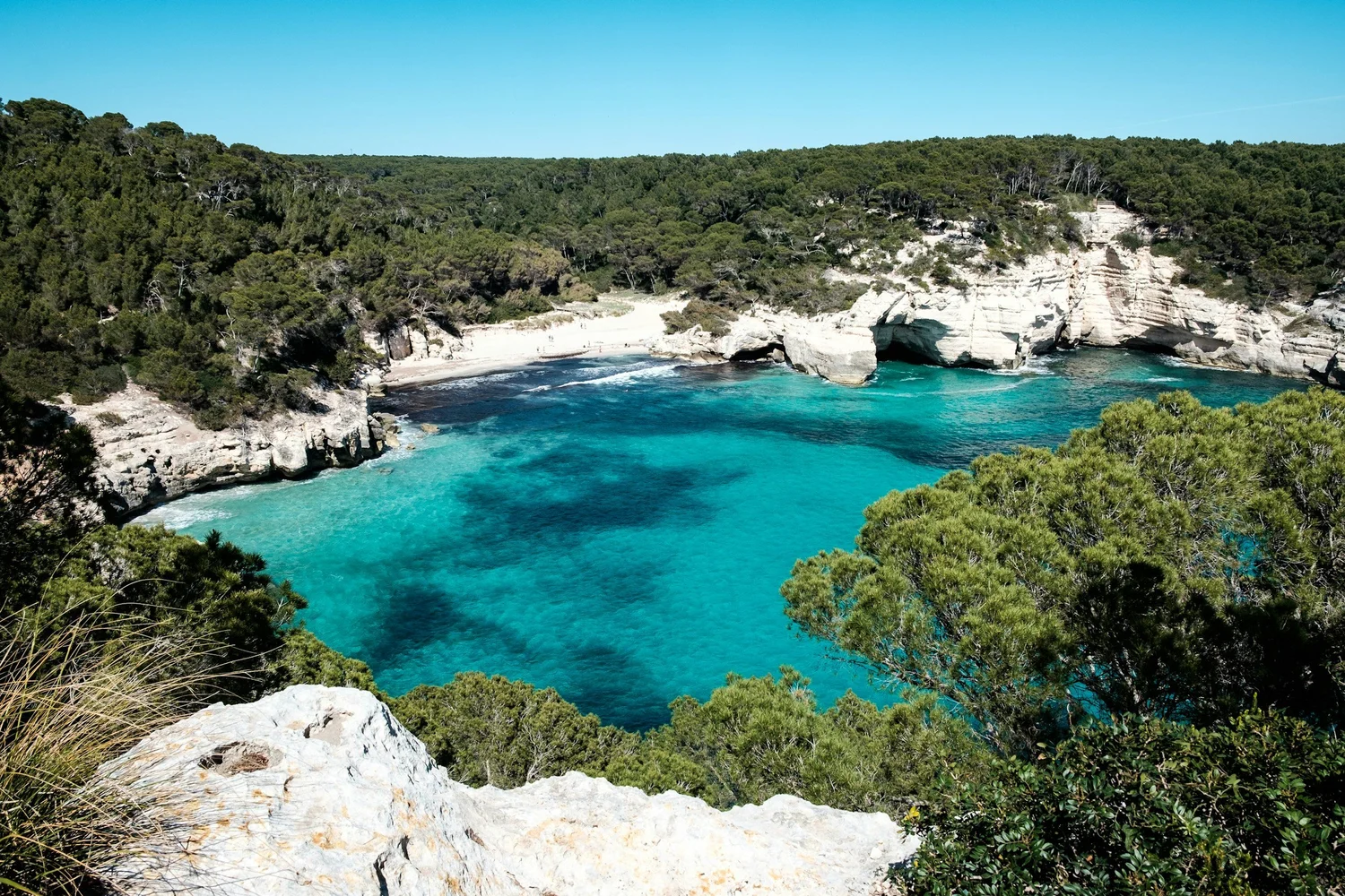 A scenic view of a coastal cove with turquoise water, white sandy beach, rocky cliffs, and dense green trees under a clear blue sky.