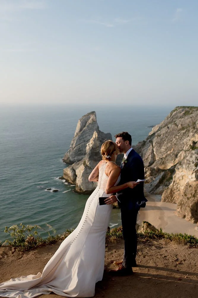 A bride and groom standing on a cliff overlooking the ocean during their wedding ceremony.