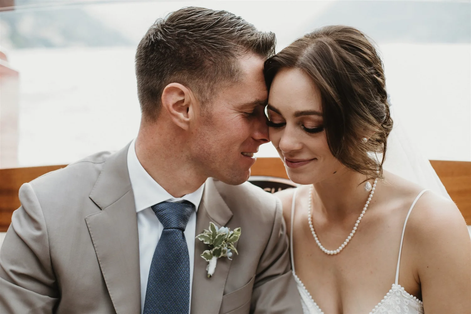 A bride and groom share an intimate moment, with foreheads touching and eyes closed, during their wedding ceremony. The groom wears a light gray suit with a white shirt and tie, while the bride is dressed in a white wedding gown with a pearl necklace