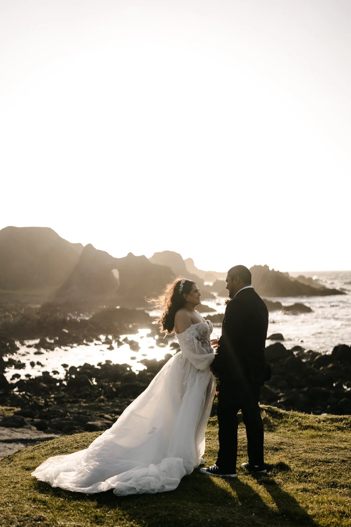 A bride and groom standing on a grassy area near the rocky shoreline of the ocean, holding hands and gazing at each other during sunset.