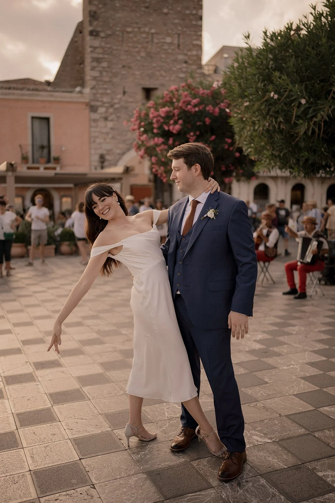A couple, possibly newlyweds, dancing together outdoors in a lively town square during sunset, with onlookers and musicians playing in the background.