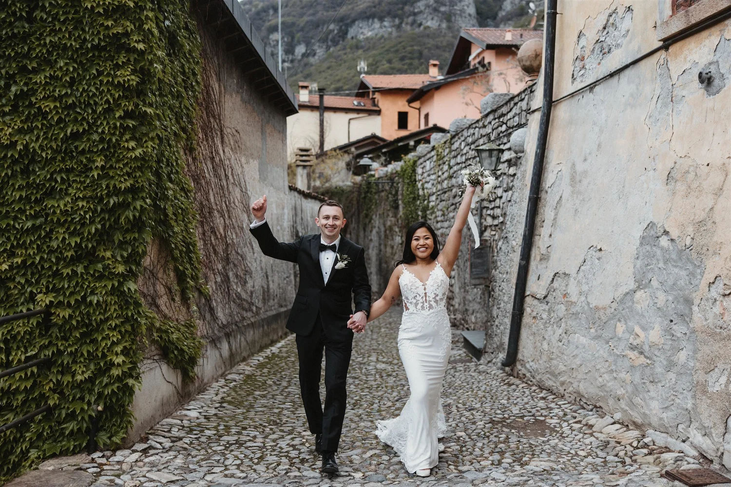A happy couple dressed in wedding attire walking hand in hand on a cobblestone street in a rustic village, smiling at the camera, with the bride holding a bouquet and raising her arm.