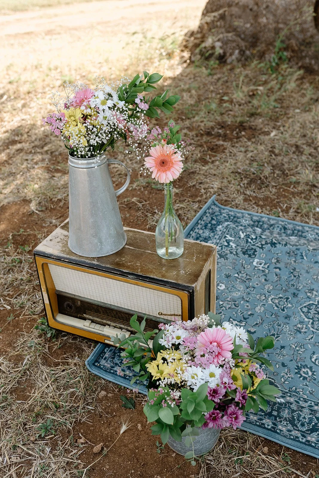 Flowers in two vases, an old radio, and a vintage rug outdoors on dirt ground.