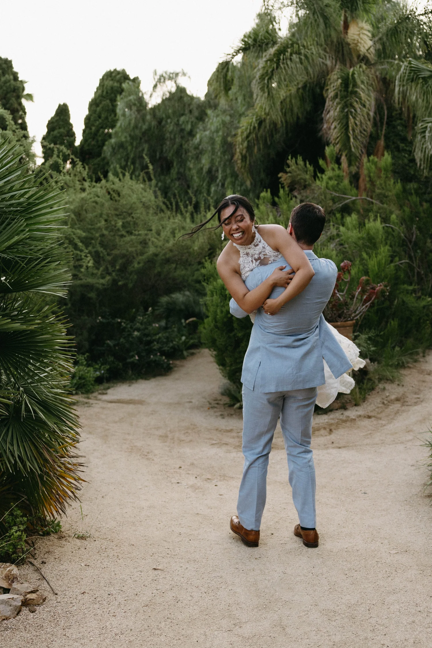 A man in a blue suit is lifting a woman in a white dress with lace detail, outdoors among lush green trees and plants, with a sandy pathway.