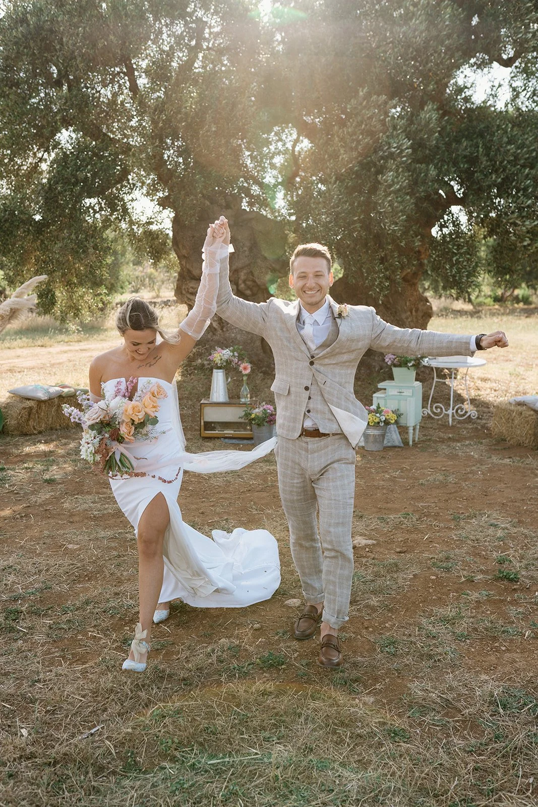A newlywed couple celebrating outdoors during sunset, with the groom in a light gray suit and the bride in a white dress holding a bouquet of flowers, smiling and raising their hands together in a joyful pose.