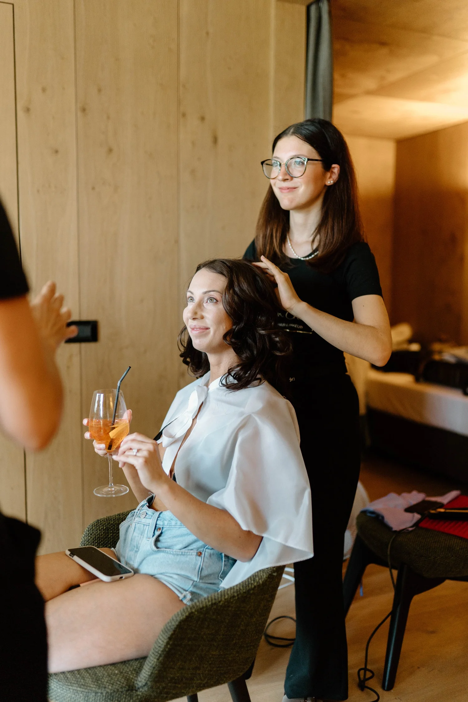 A woman with long dark hair, wearing a white shirt and denim shorts, is sitting at a table holding a glass of an orange cocktail with a straw. A makeup artist with glasses and dark hair is standing behind her, styling her hair in a casual indoor sett