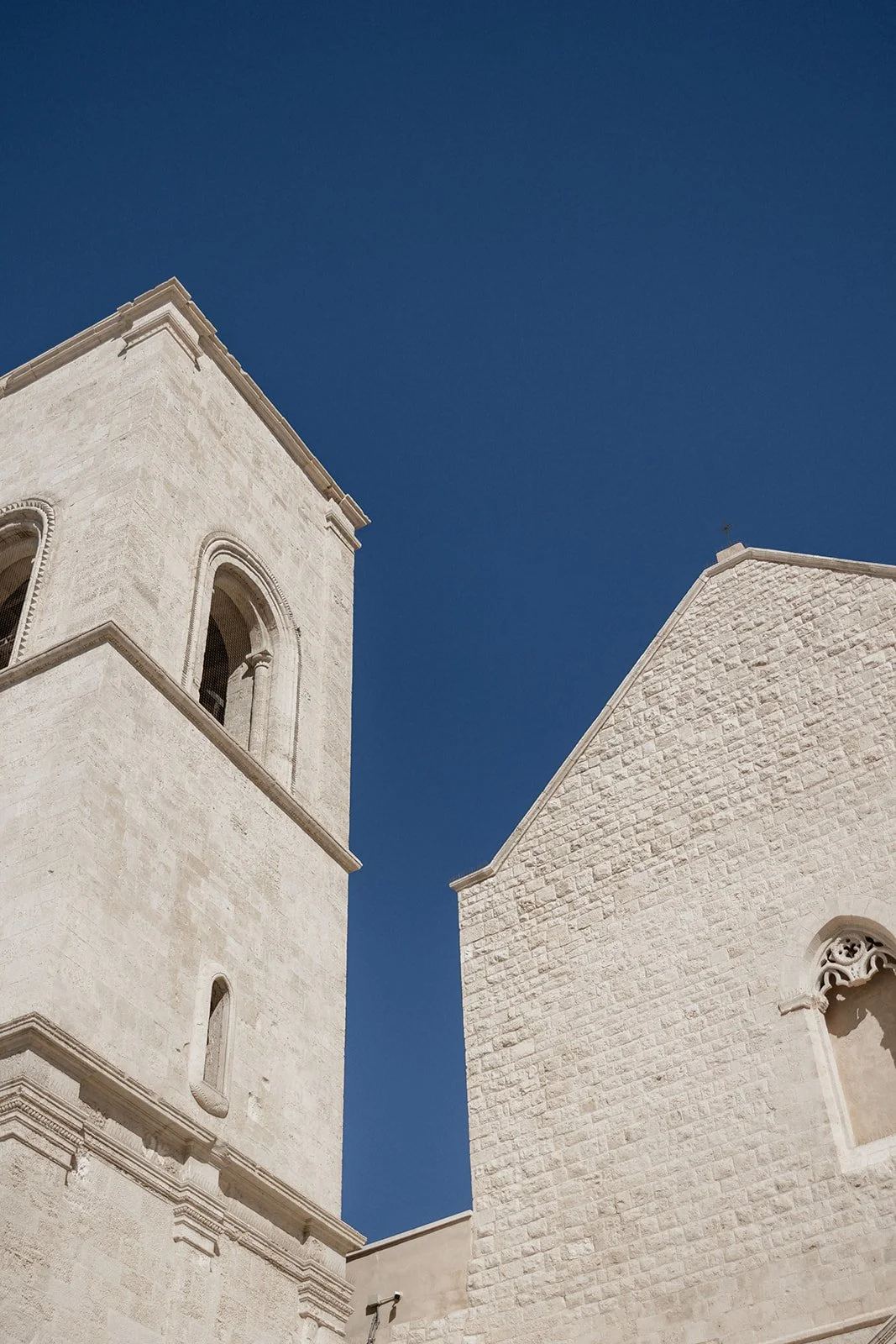 White stone church with a bell tower on the left and a textured wall on the right, against a clear blue sky.