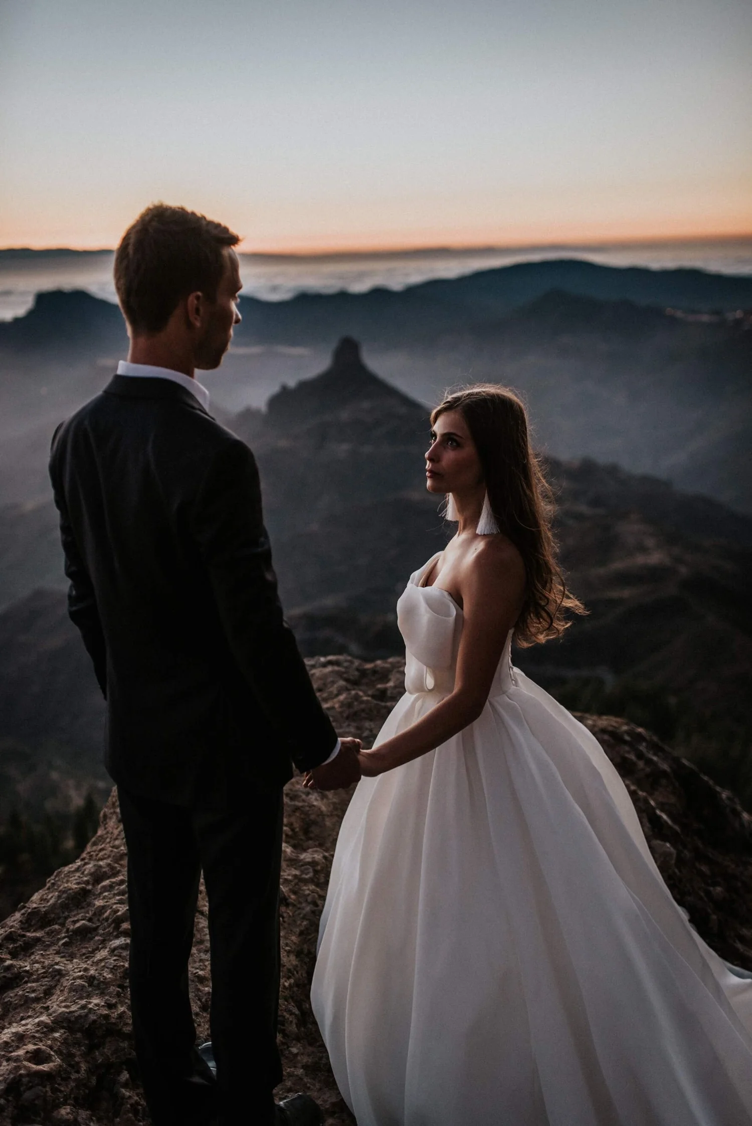 A man and woman holding hands on a rocky landscape with mountains and a sunset sky in the background.
