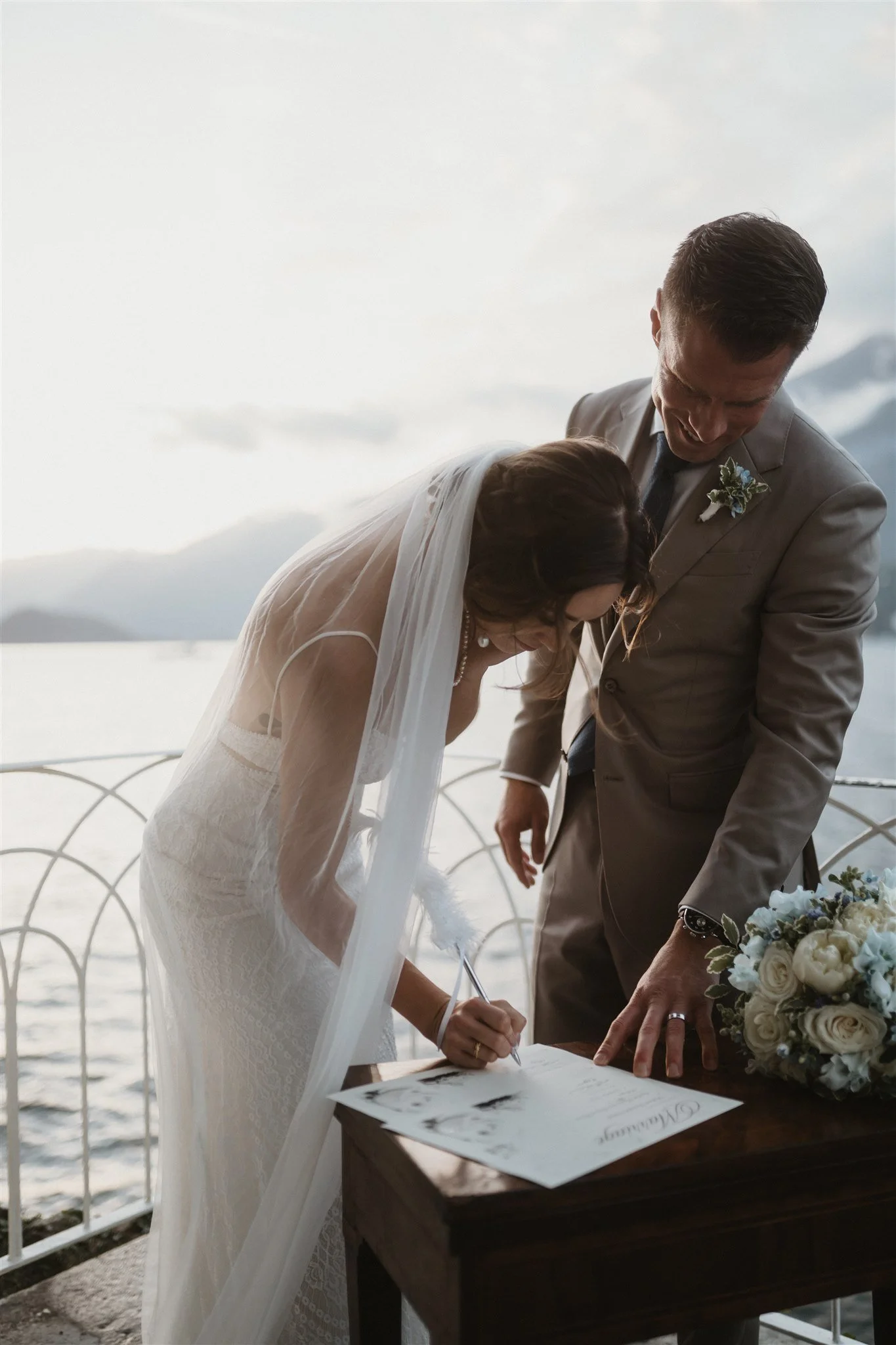 A bride and groom signing a marriage license outdoors by a body of water with mountains in the background.