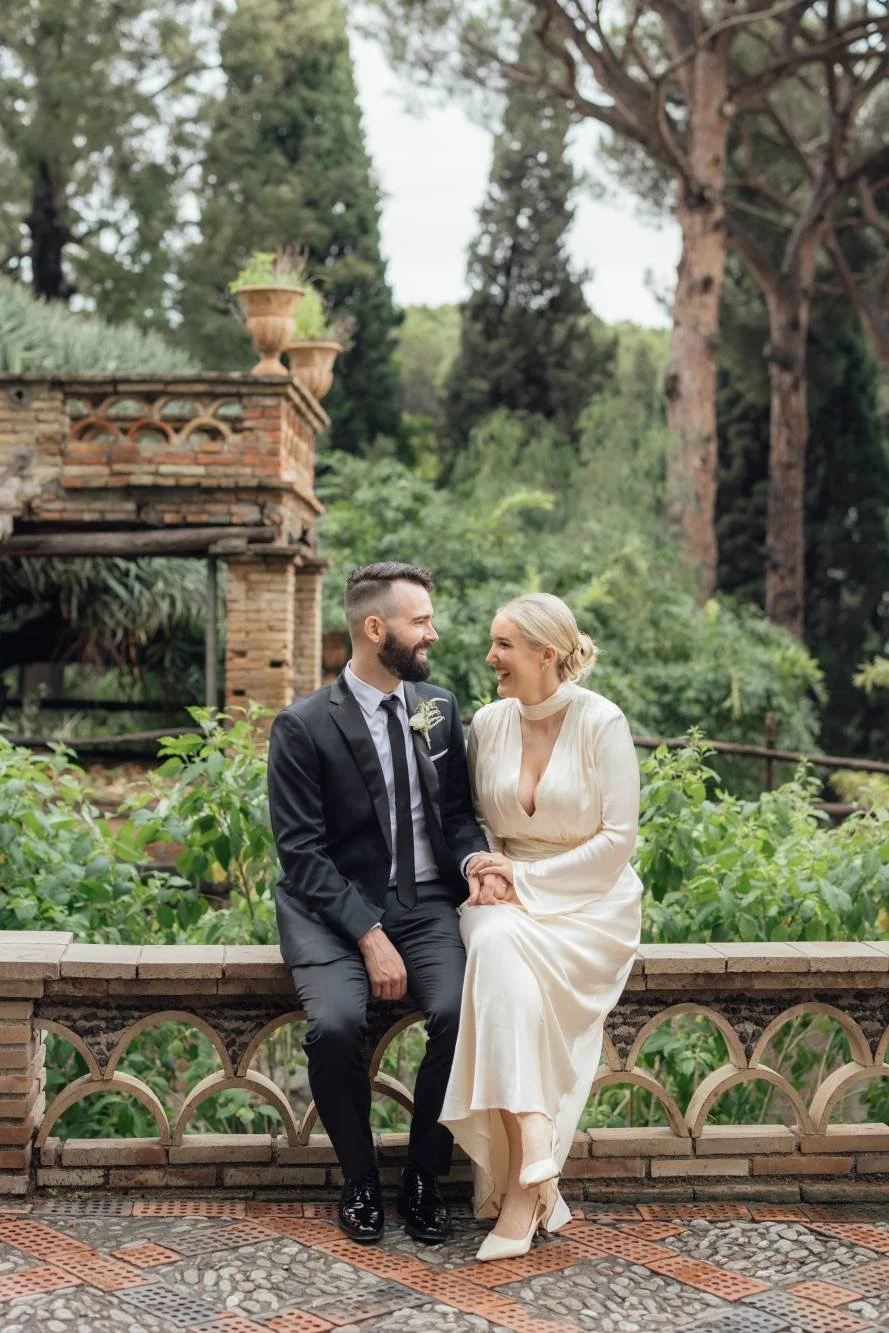 A bride and groom sitting closely together outdoors, holding hands, and sharing a joyful moment. The bride wears a cream-colored dress with a plunging neckline and a choker, and the groom is in a black suit with a tie. They are surrounded by lush greenery and tall trees.