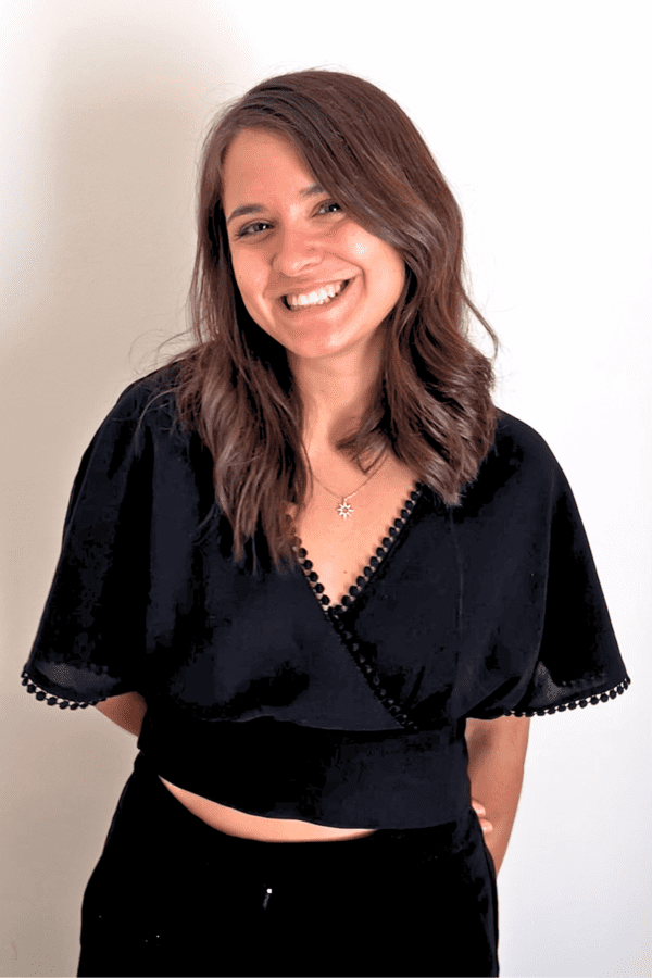 Woman with wavy brown hair smiling, wearing a black top with scalloped trim and a star-shaped necklace, standing against a light background.