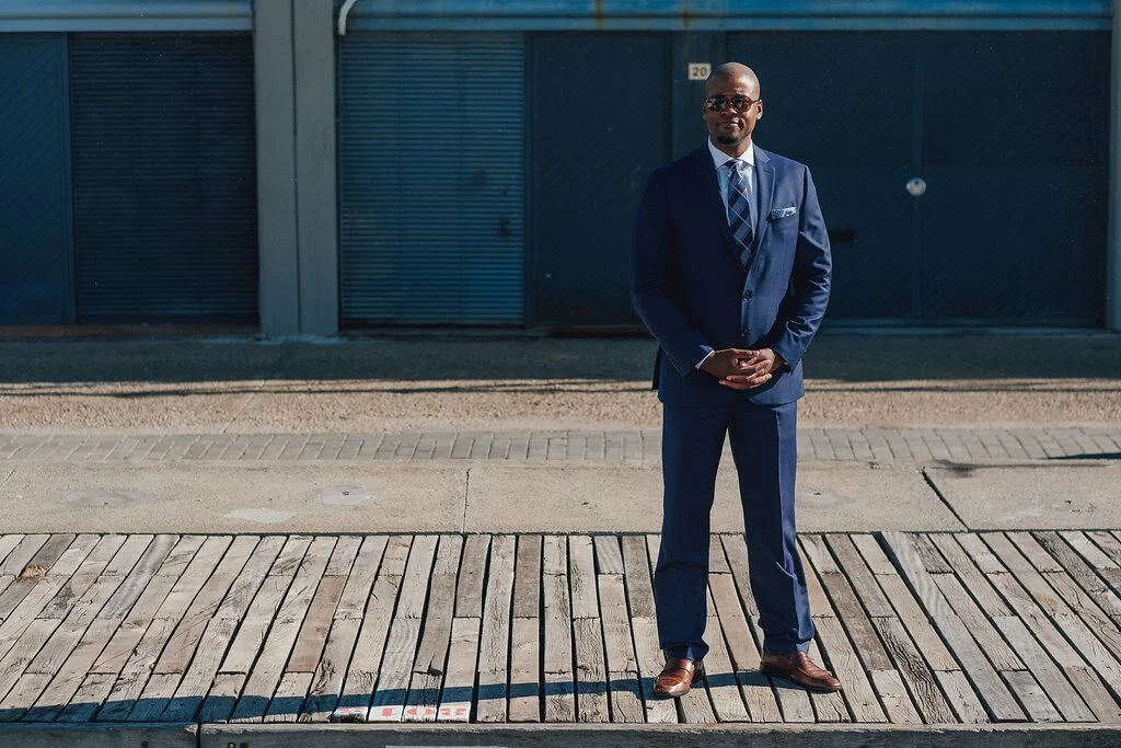 A man in a blue suit and sunglasses standing outdoors with his hands clasped in front of him.
