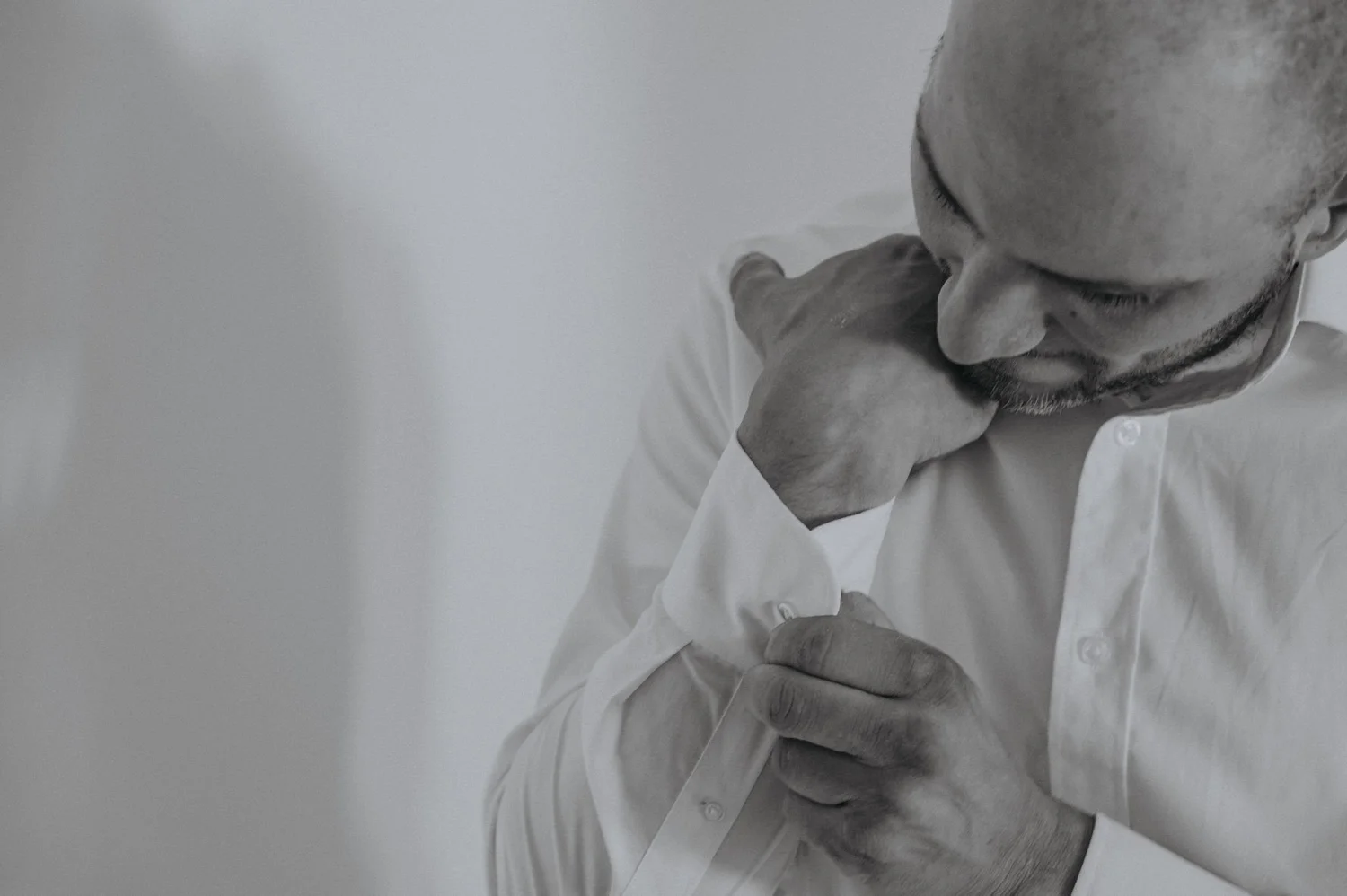 A man with a shaved head and beard wearing a white shirt, resting his chin on his hand, looking down, against a plain white background.