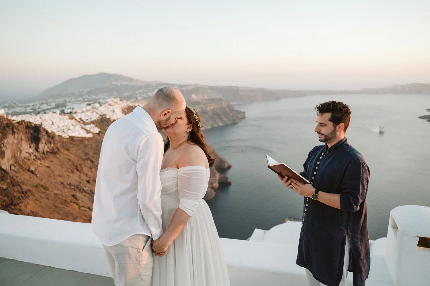 A couple exchanging wedding vows by the water on a cliffside during sunset, with a man officiating.