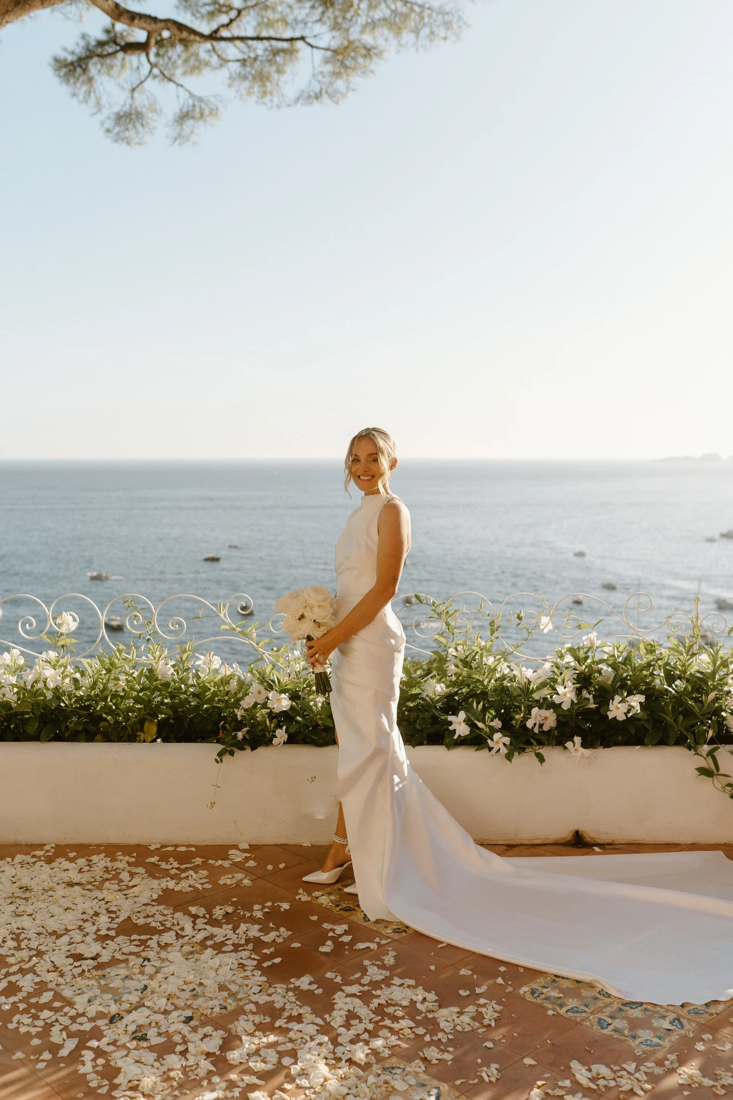 A smiling bride in a white wedding dress holding a bouquet of white flowers stands on a terrace overlooking the ocean. There are white flower petals scattered on the ground and white flowers along the terrace railing. A tree is visible in the backgro