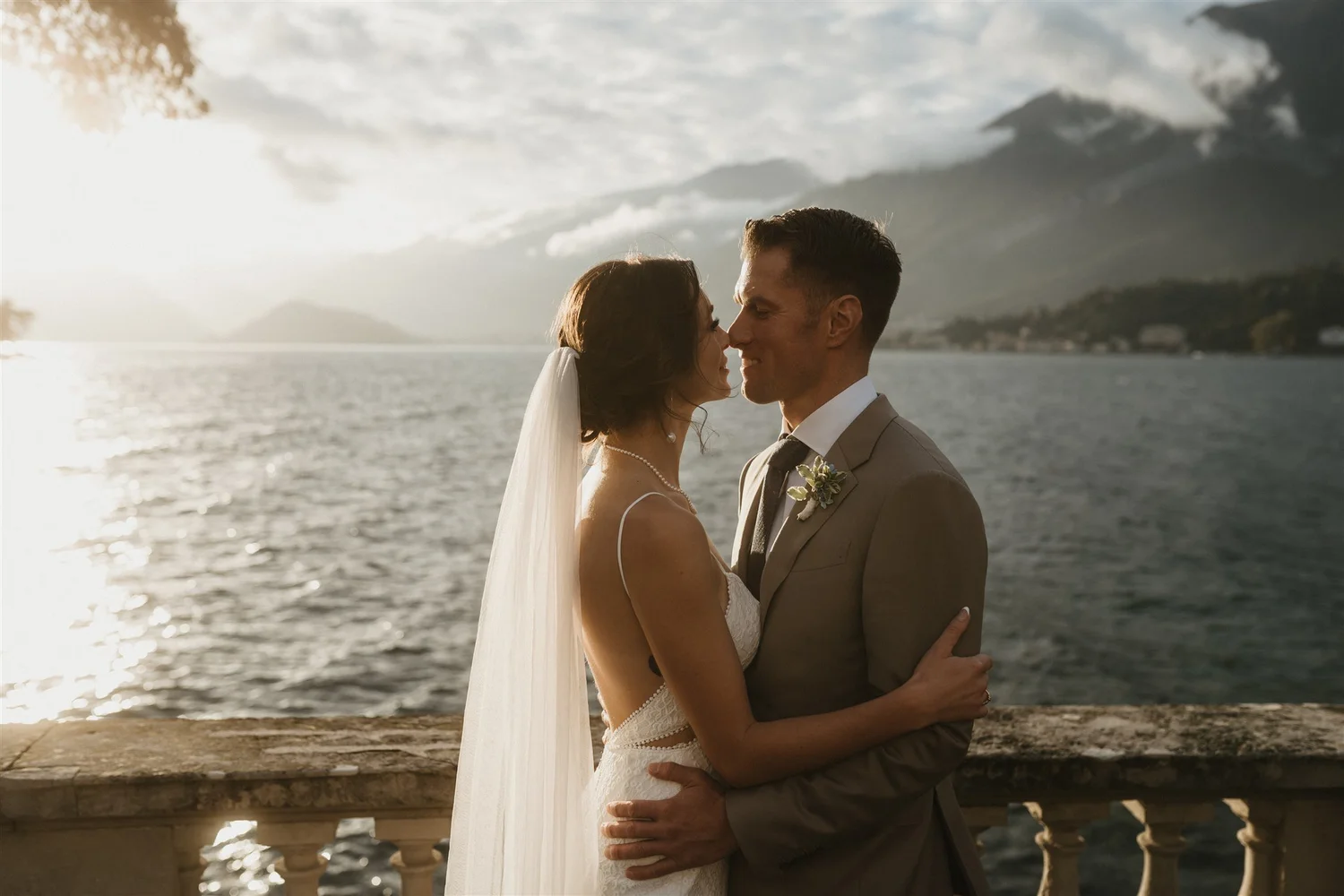 A bride and groom embracing near a body of water during sunset, with mountains in the background.